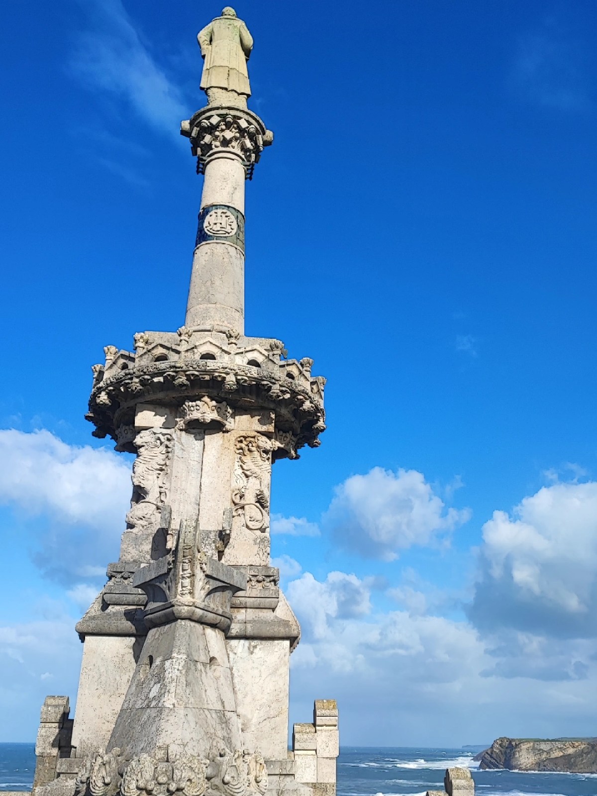 Stone monument with a statue on top near the ocean under a blue sky.