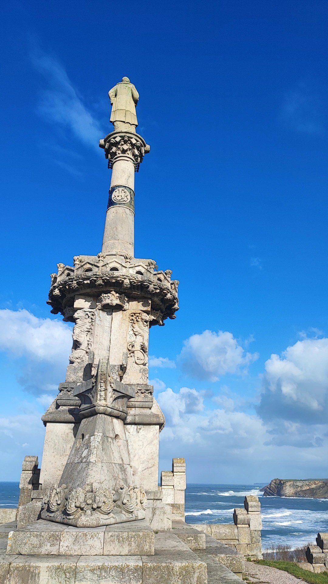 Stone monument with a statue on top near the ocean under a blue sky.