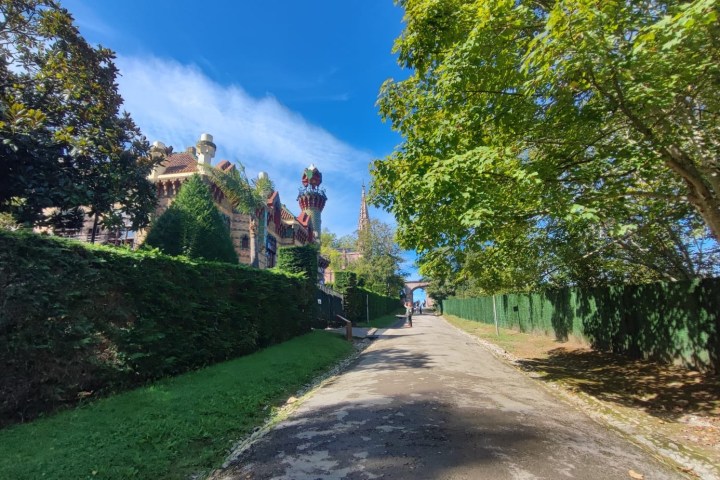 Path with green hedges and trees leading to a colorful, ornate building under a clear blue sky.
