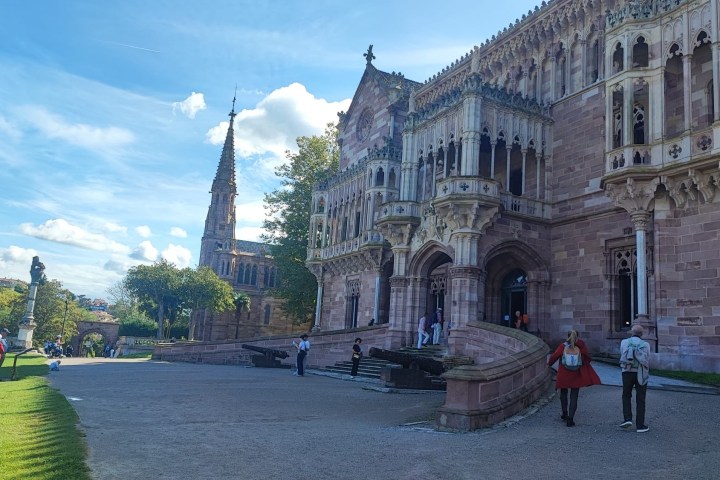 People walking by a historic building with ornate architecture on a sunny day.