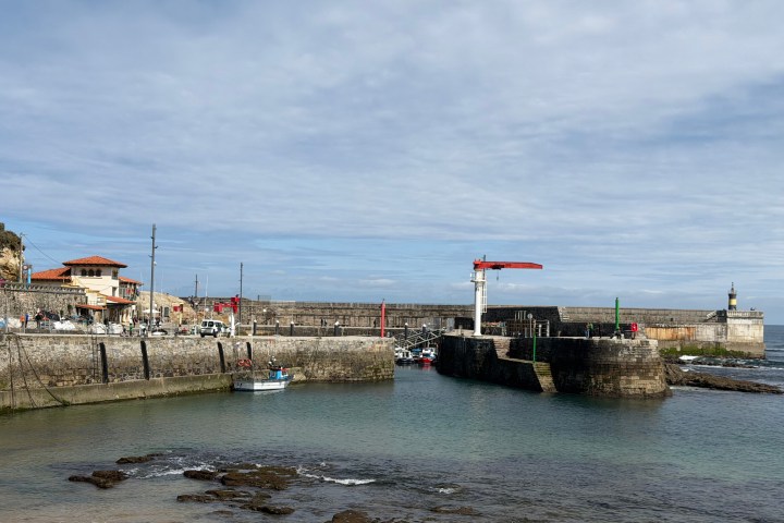 Harbor scene with lighthouse and boats docked along stone walls under a cloudy sky.