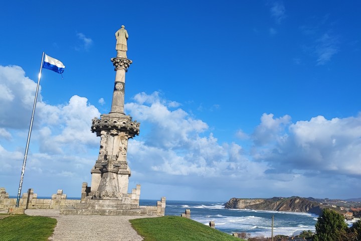 Stone monument and flagpole by the sea under a blue sky with clouds.