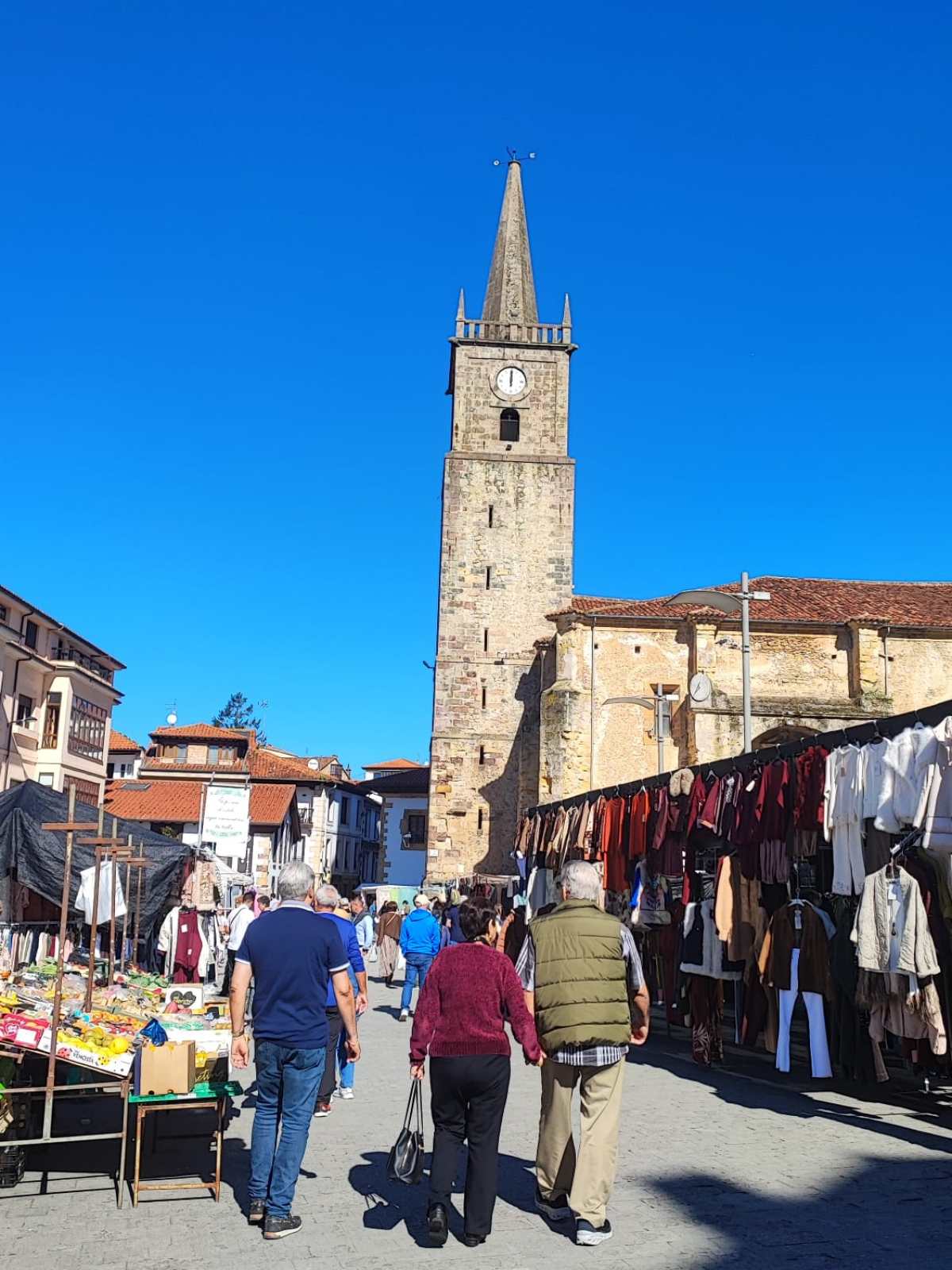 People walking in a market with a tall clock tower in the background under clear blue sky.