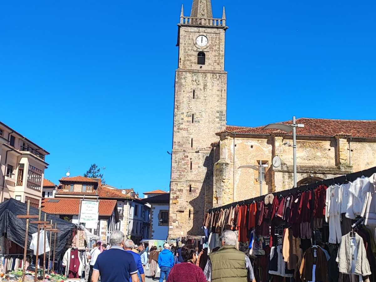 People walking in a market with a tall clock tower in the background under clear blue sky.