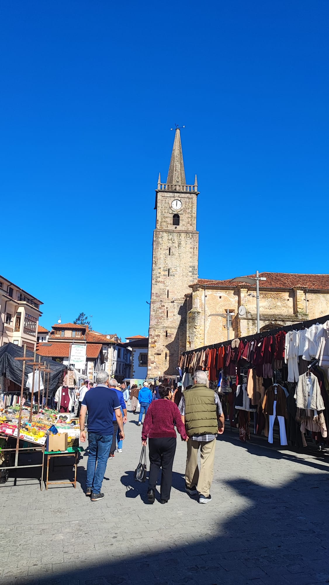 People walking in a market with a tall clock tower in the background under clear blue sky.