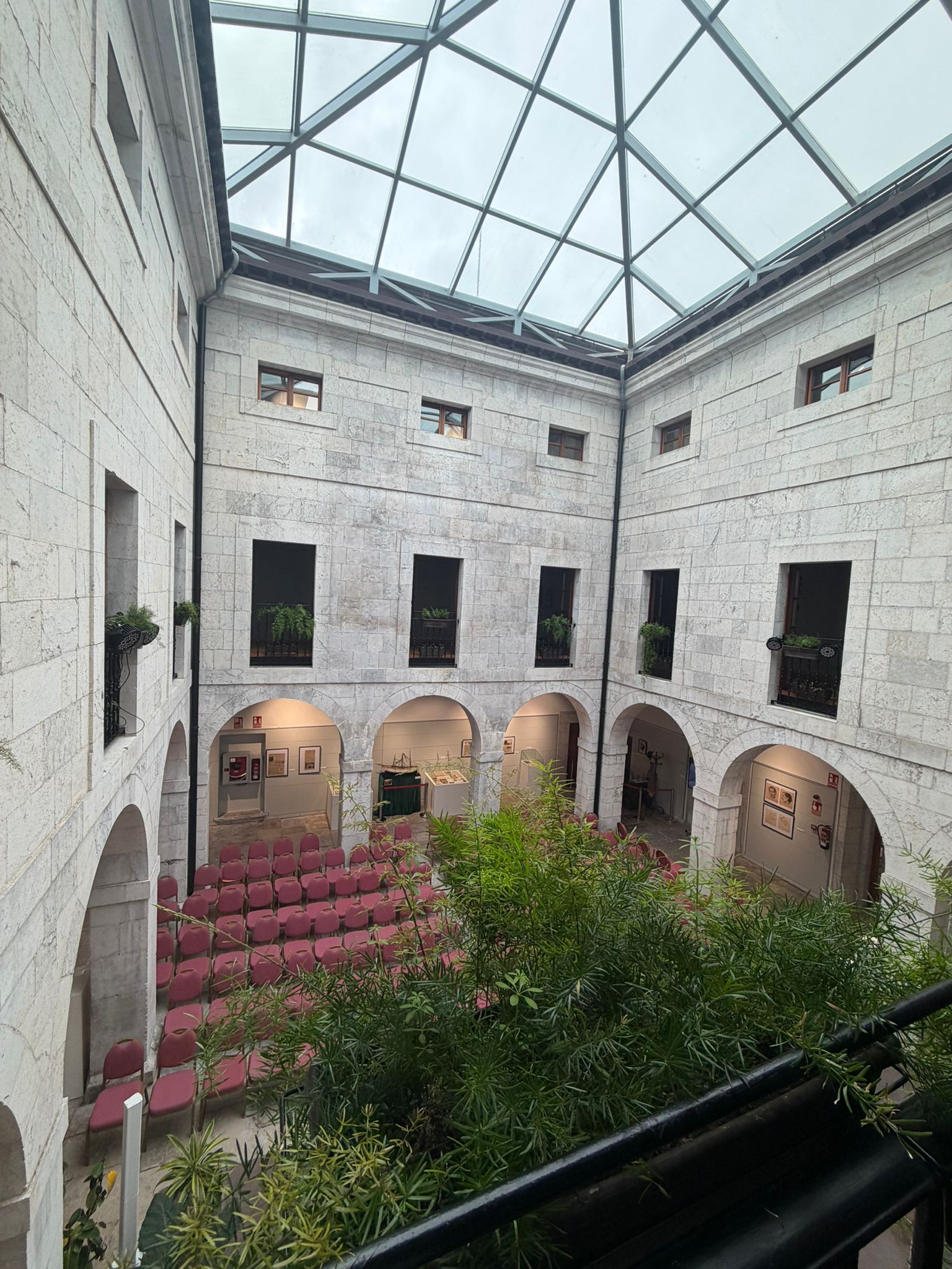Courtyard with stone walls, red chairs, plants, and large glass ceiling.