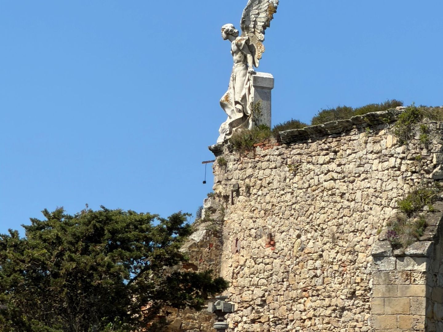 Stone angel statue atop a historic stone wall with trees below, under a clear blue sky.