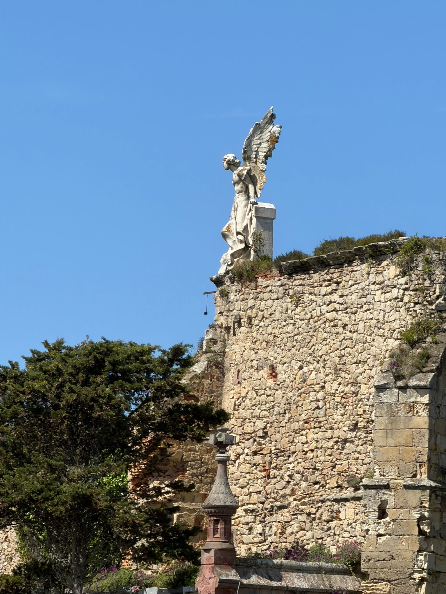 Stone angel statue atop a historic stone wall with trees below, under a clear blue sky.