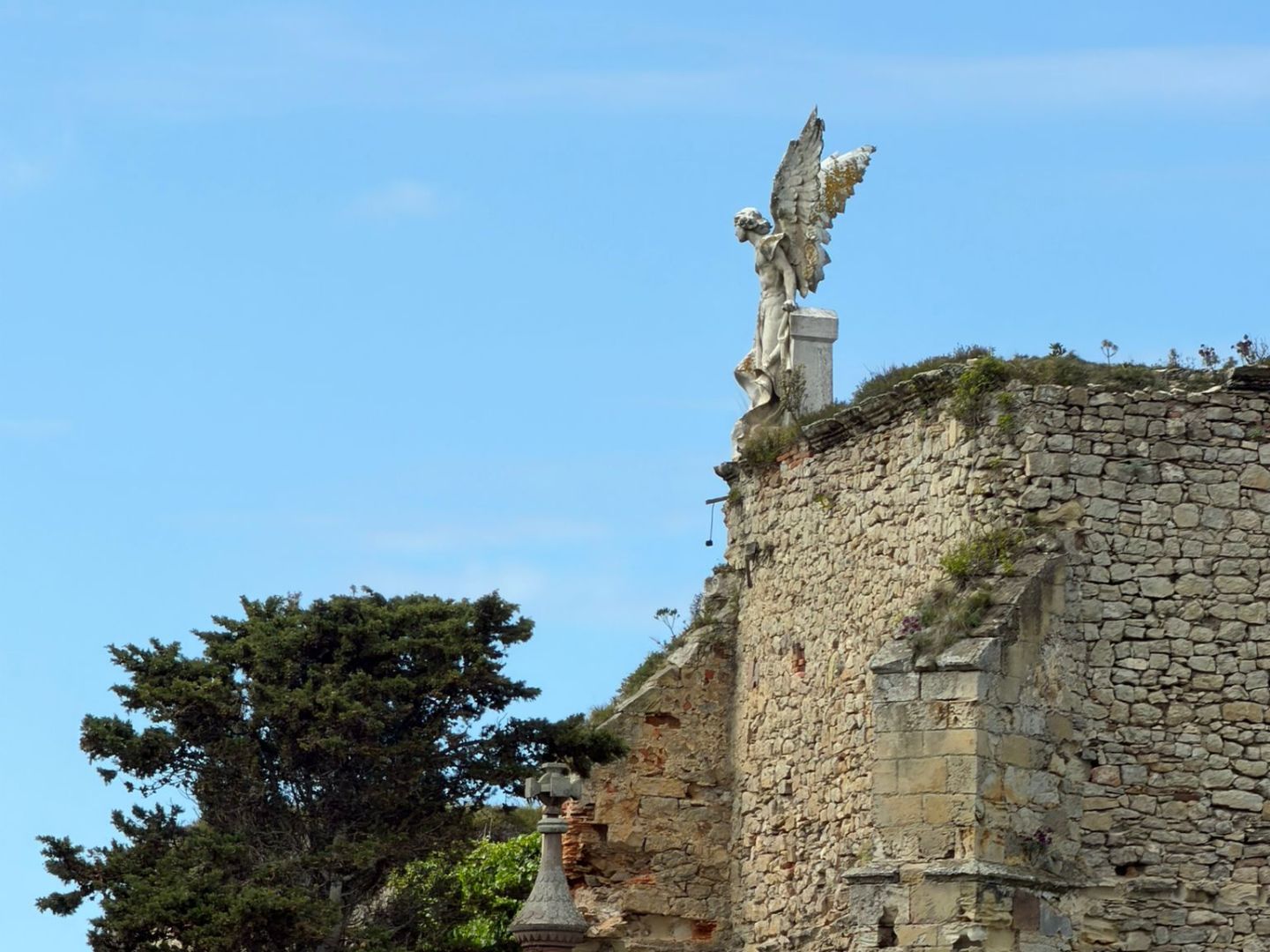 Stone wall with a winged statue and trees against a blue sky.