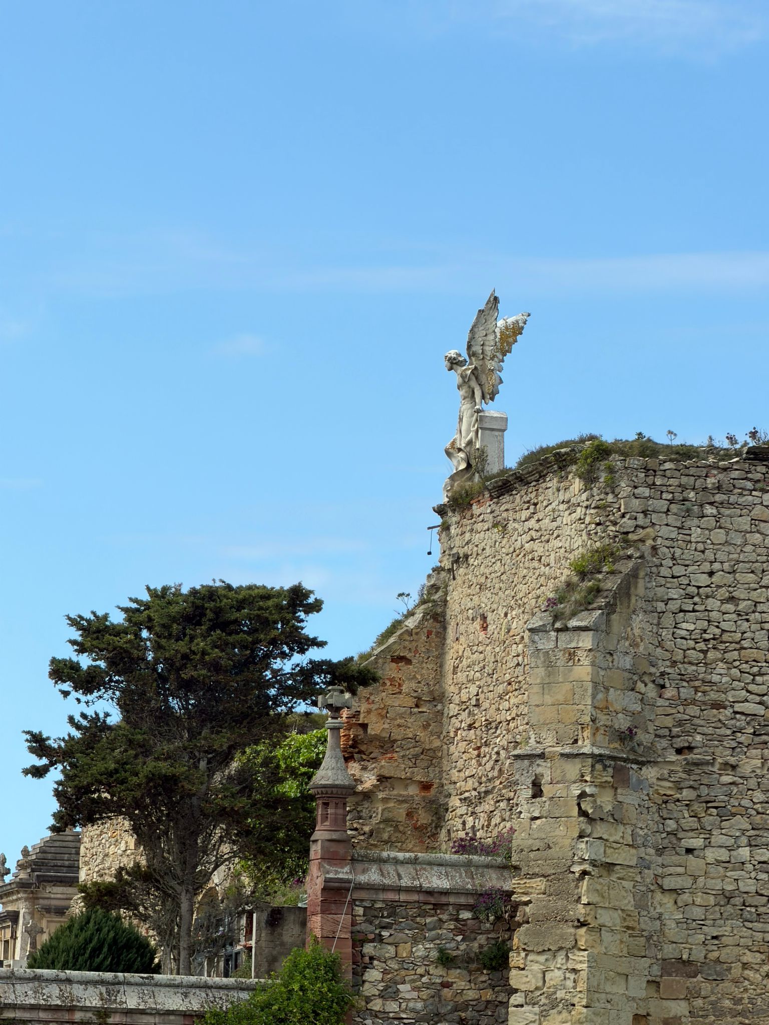 Stone wall with a winged statue and trees against a blue sky.