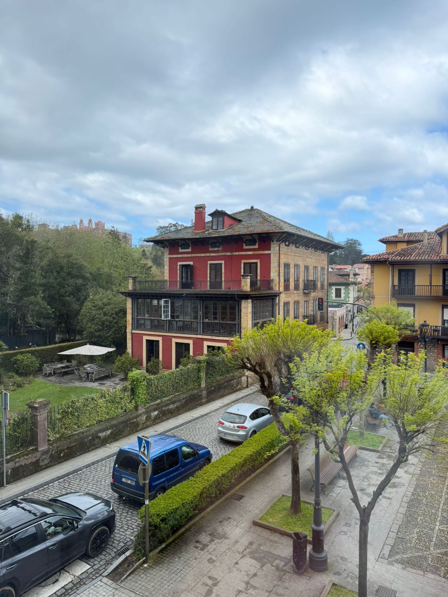 Street view with colorful buildings, parked cars, and trees under a cloudy sky.