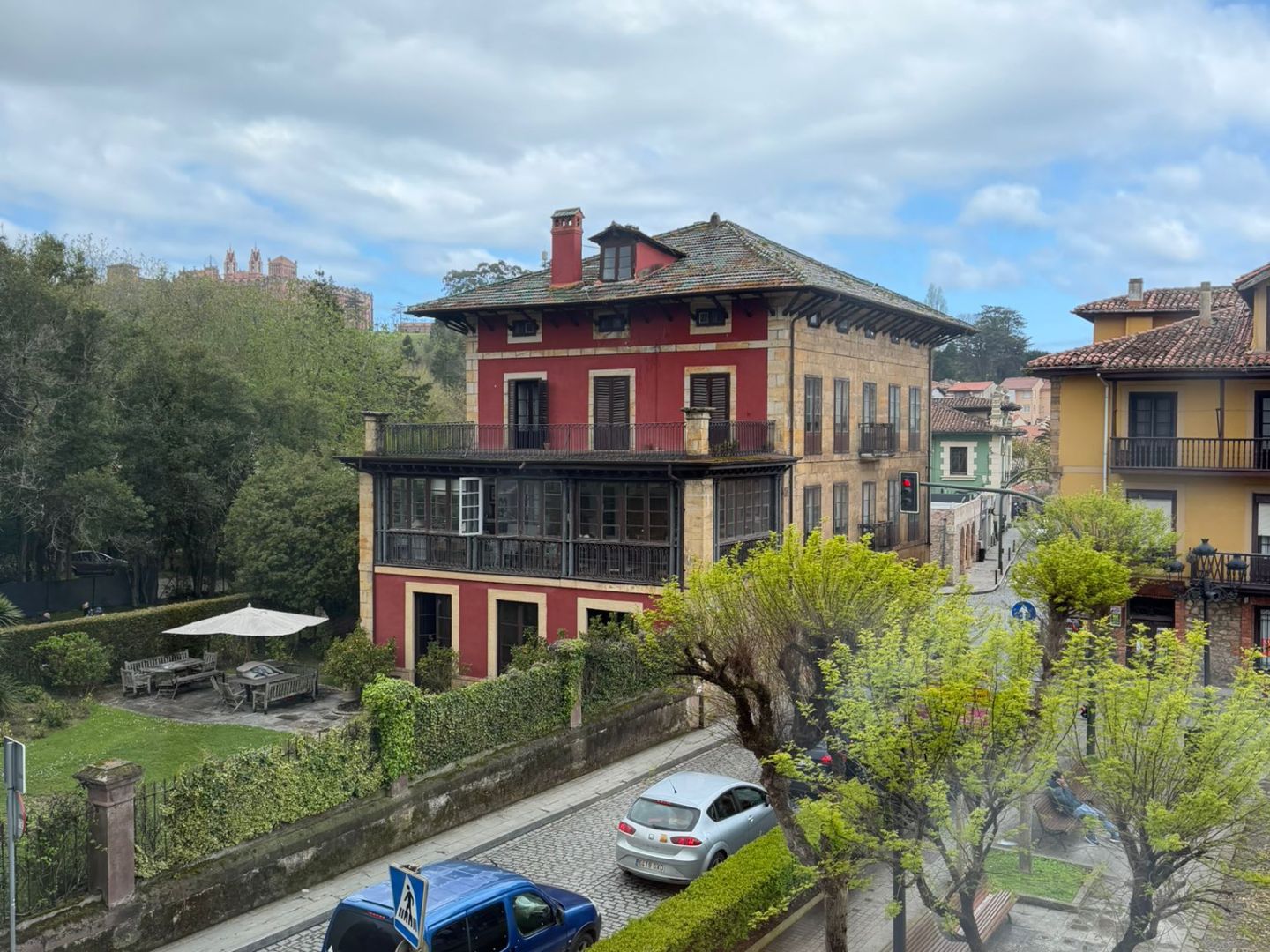 Street view with colorful buildings, parked cars, and trees under a cloudy sky.