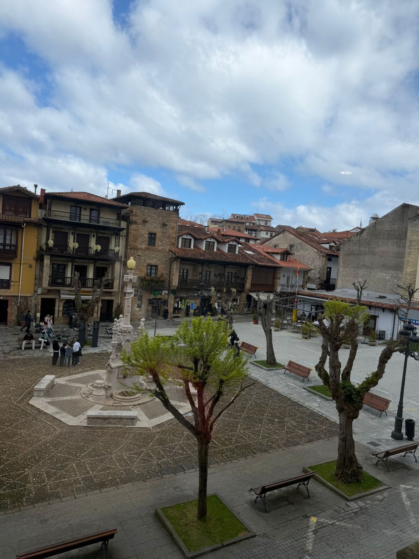 Open plaza with trees, benches, a stone fountain, and surrounding old buildings under a cloudy sky.
