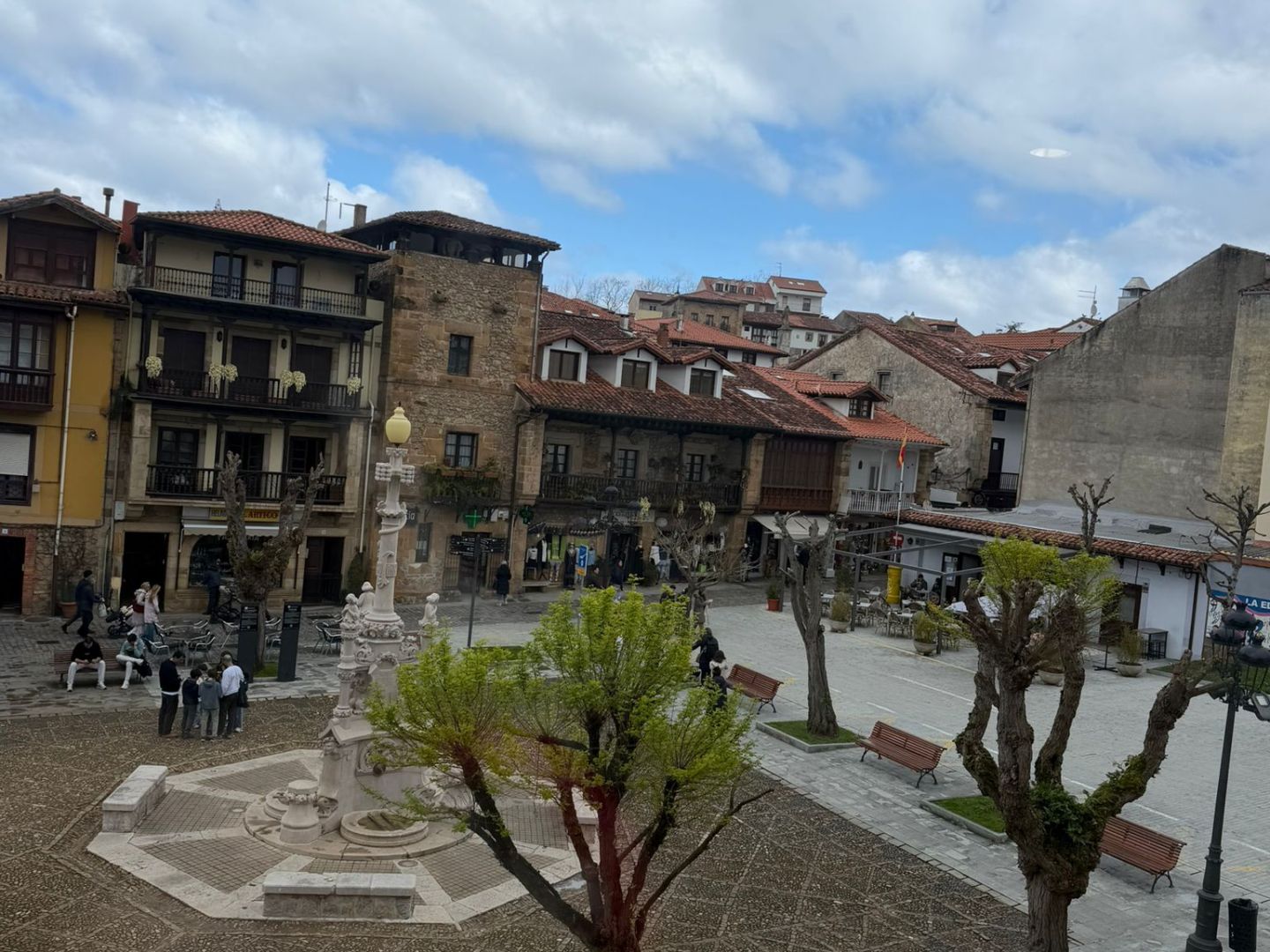 Open plaza with trees, benches, a stone fountain, and surrounding old buildings under a cloudy sky.