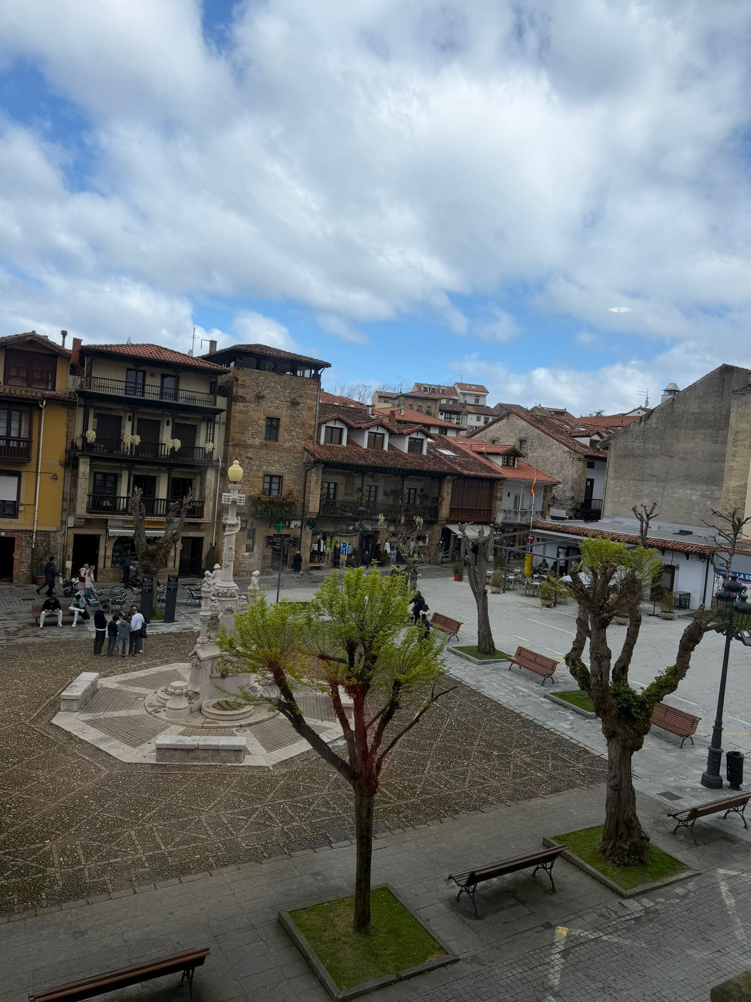 Open plaza with trees, benches, a stone fountain, and surrounding old buildings under a cloudy sky.