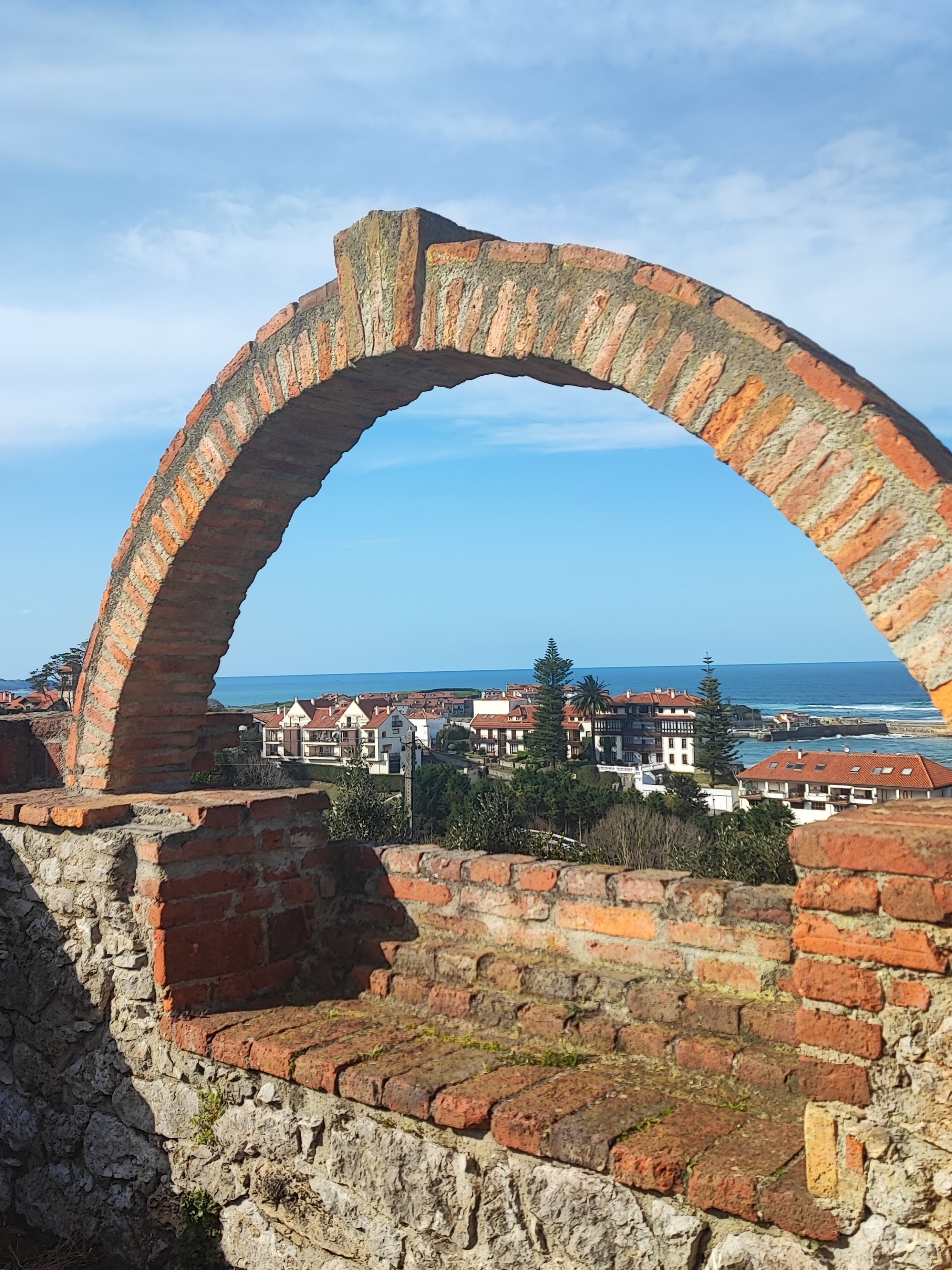 Stone arch overlooking a coastal town with blue sky.