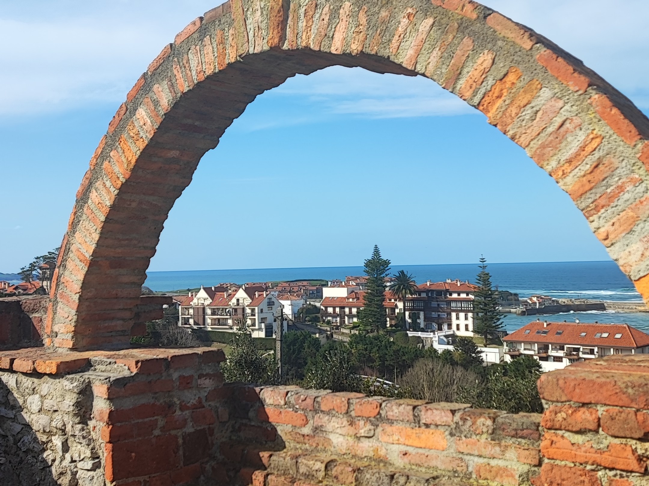 Stone arch overlooking a coastal town with blue sky.