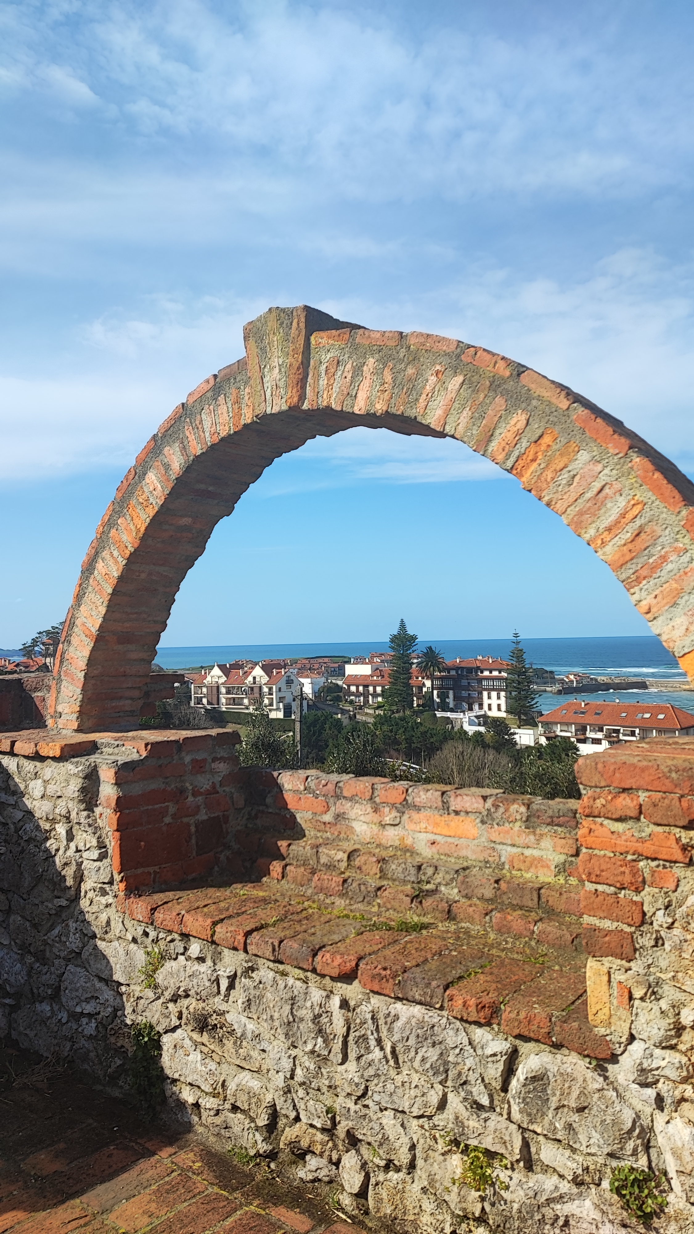 Stone arch overlooking a coastal town with blue sky.