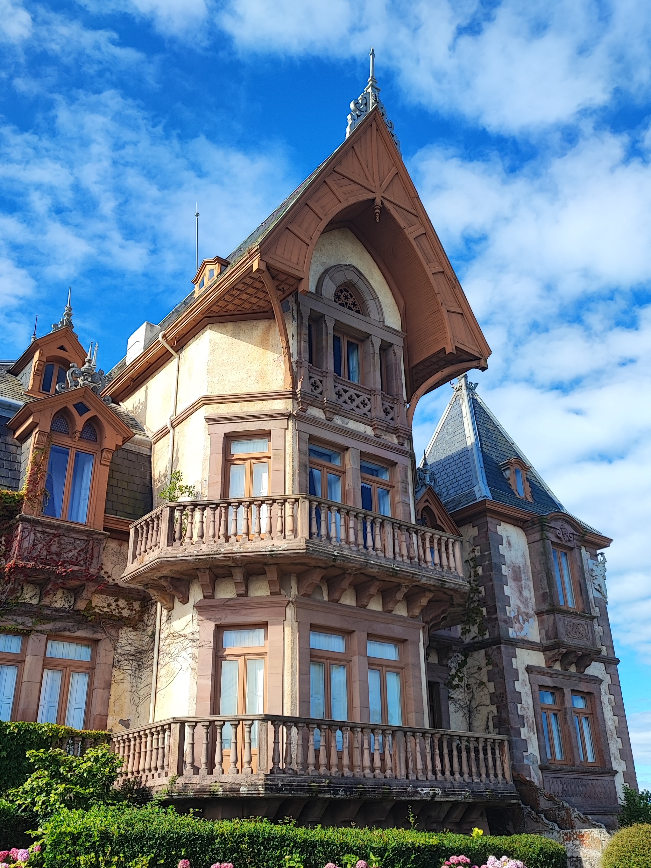 Historic mansion with ornate balcony, pointed roof, and blooming hydrangeas under a blue sky.