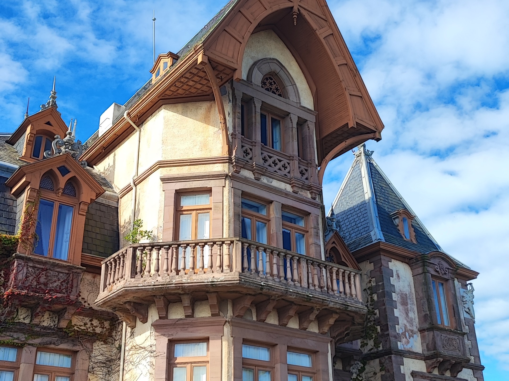 Historic mansion with ornate balcony, pointed roof, and blooming hydrangeas under a blue sky.