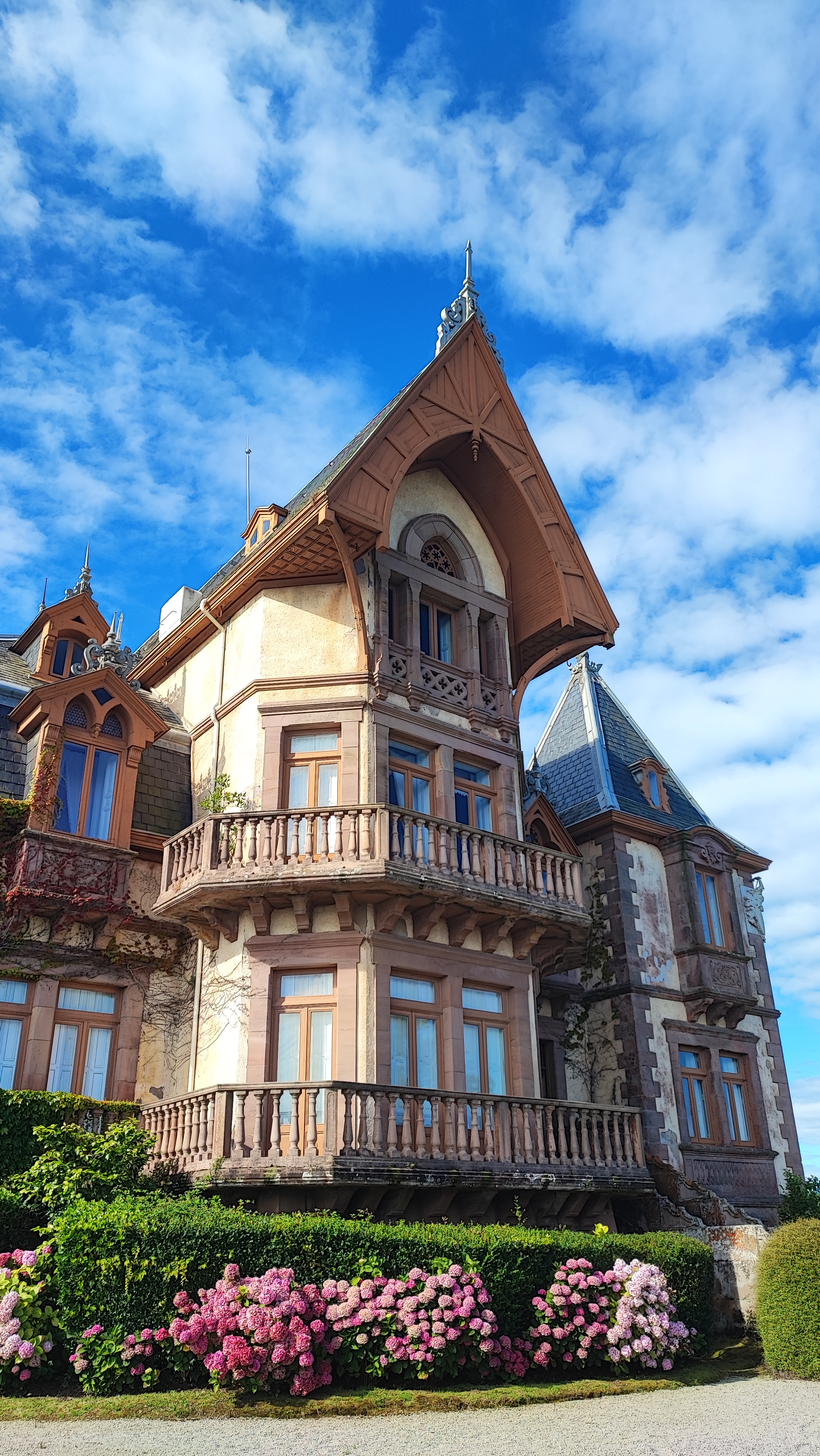 Historic mansion with ornate balcony, pointed roof, and blooming hydrangeas under a blue sky.