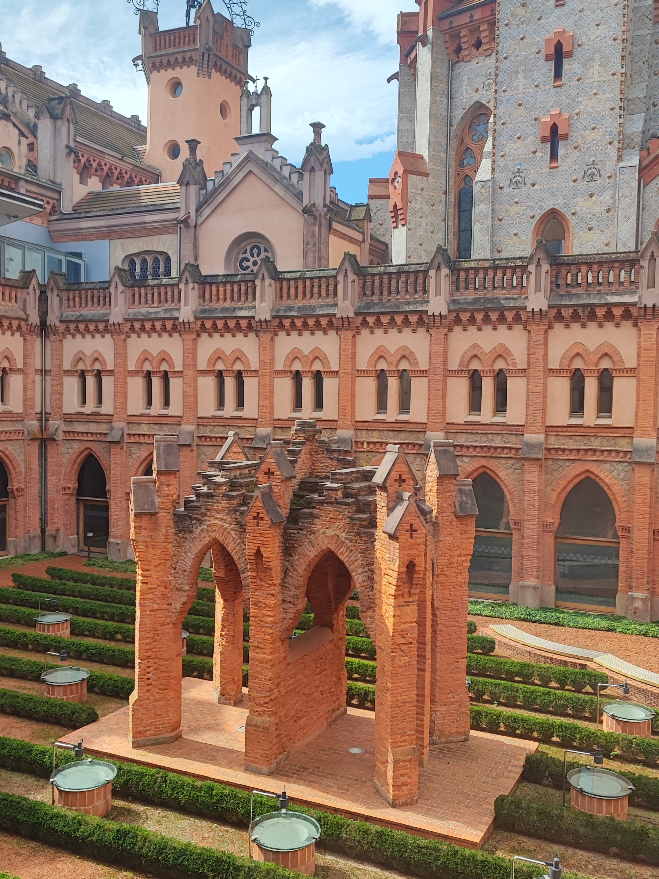 Historic brick courtyard with arches, hedges, and a tall tower in the background.