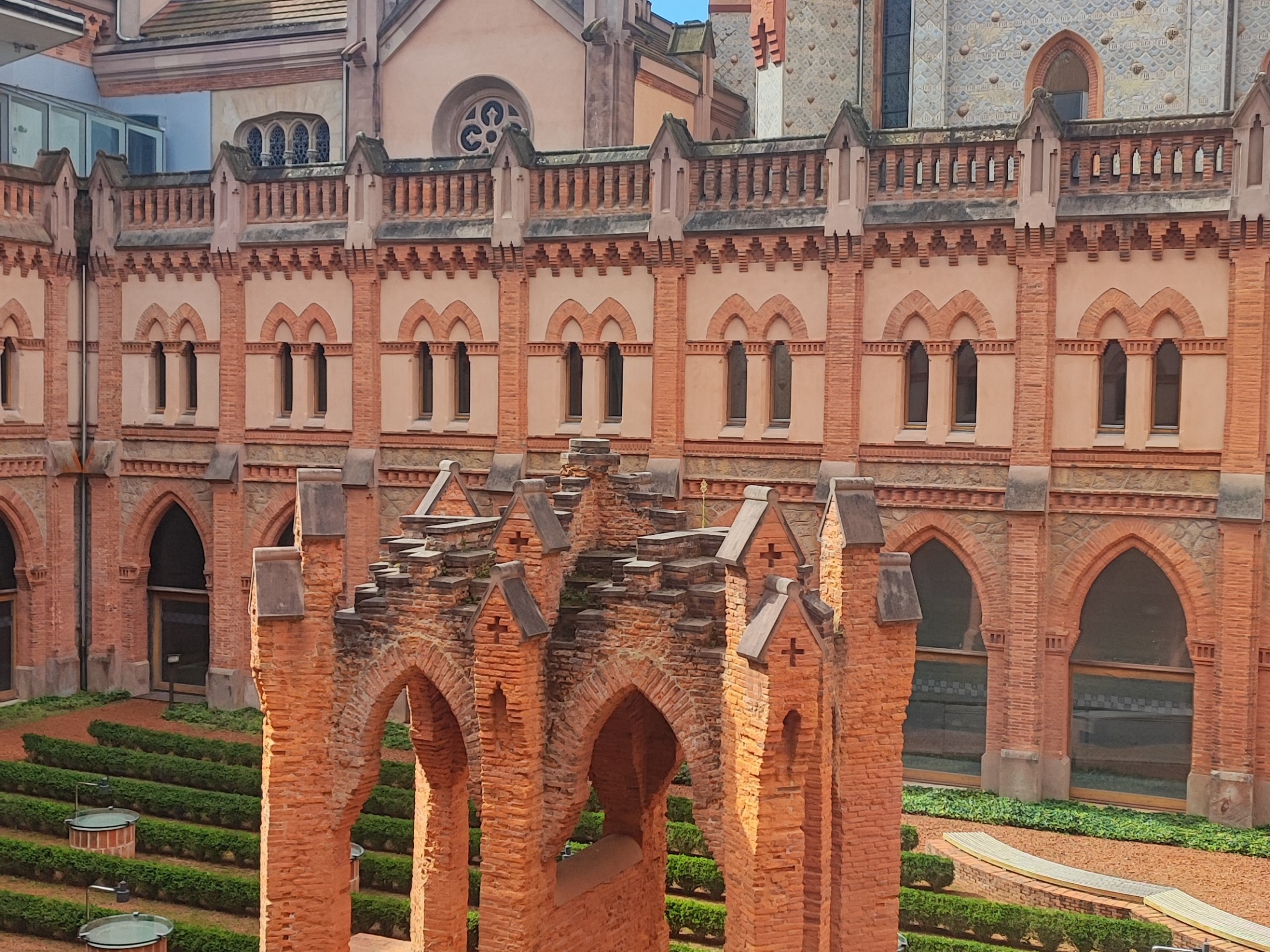 Historic brick courtyard with arches, hedges, and a tall tower in the background.