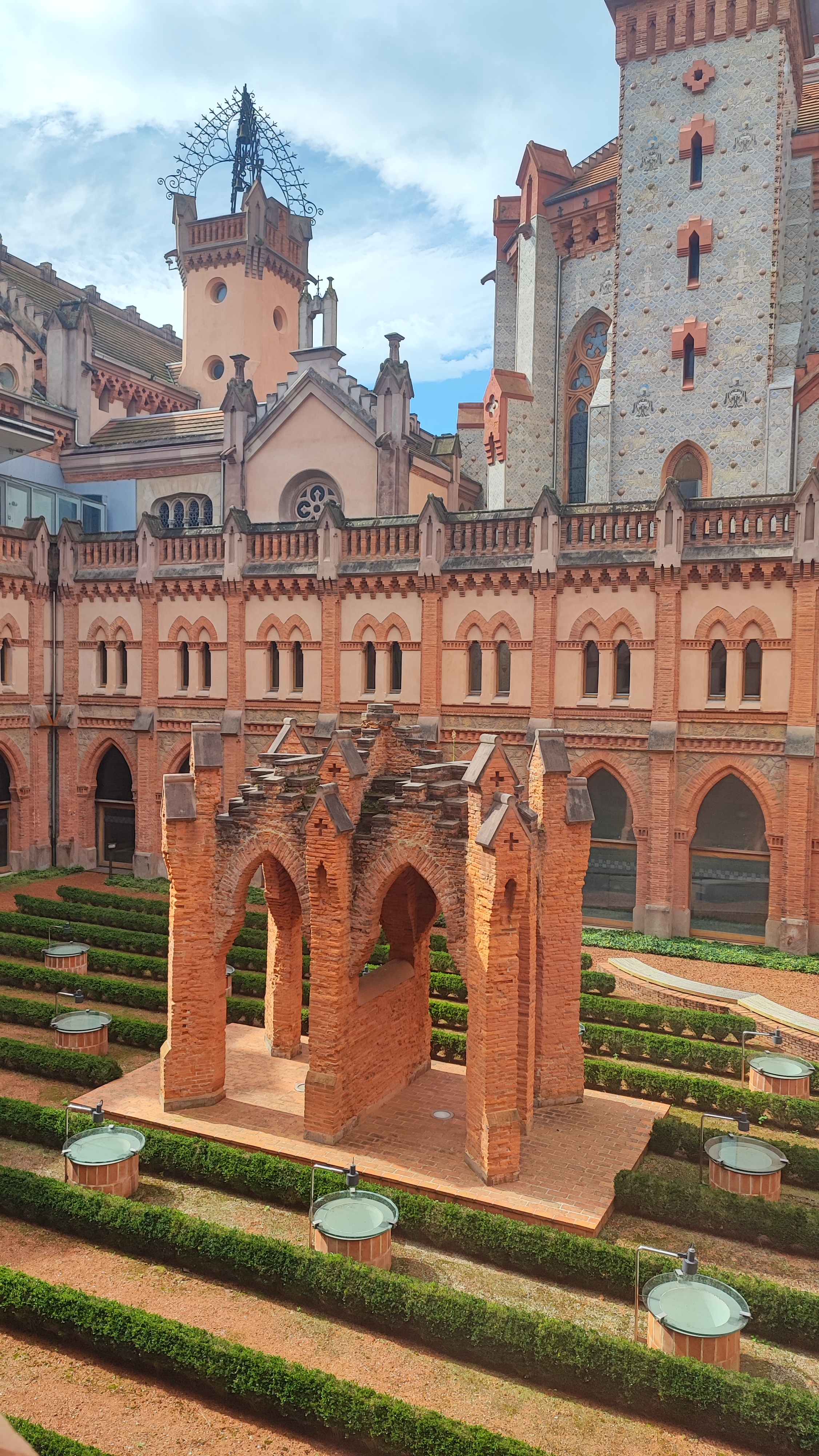 Historic brick courtyard with arches, hedges, and a tall tower in the background.