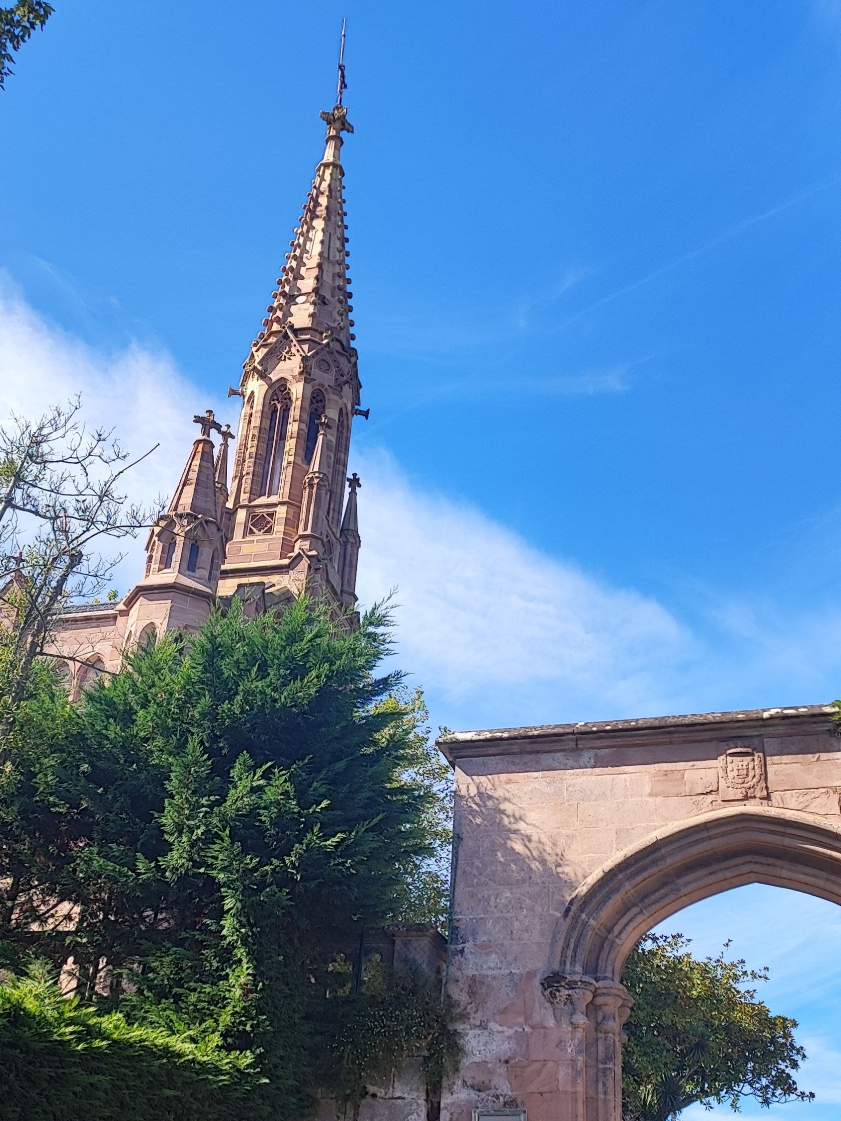 Gothic-style church spire with trees and archway under a blue sky.