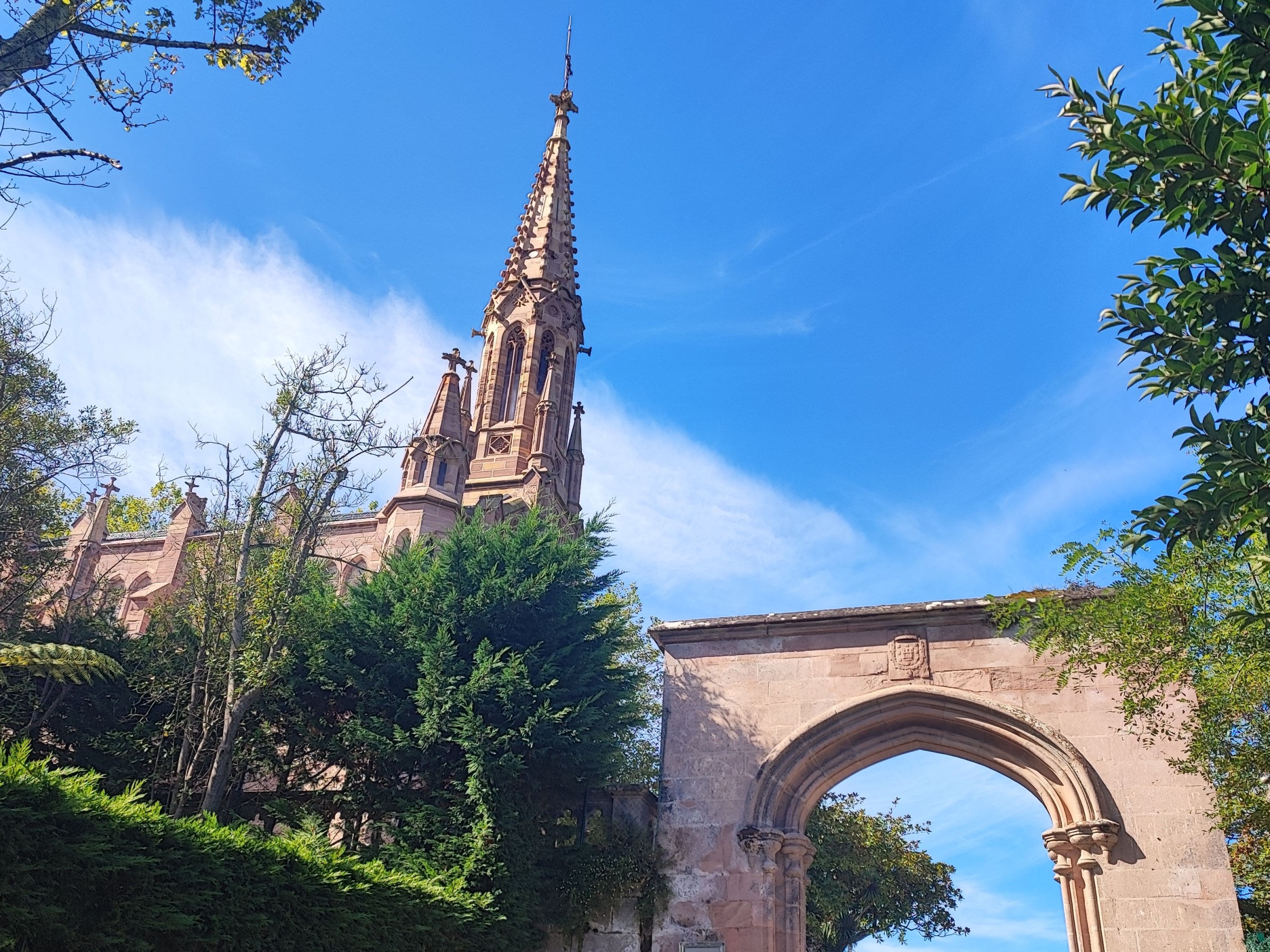 Gothic-style church spire with trees and archway under a blue sky.