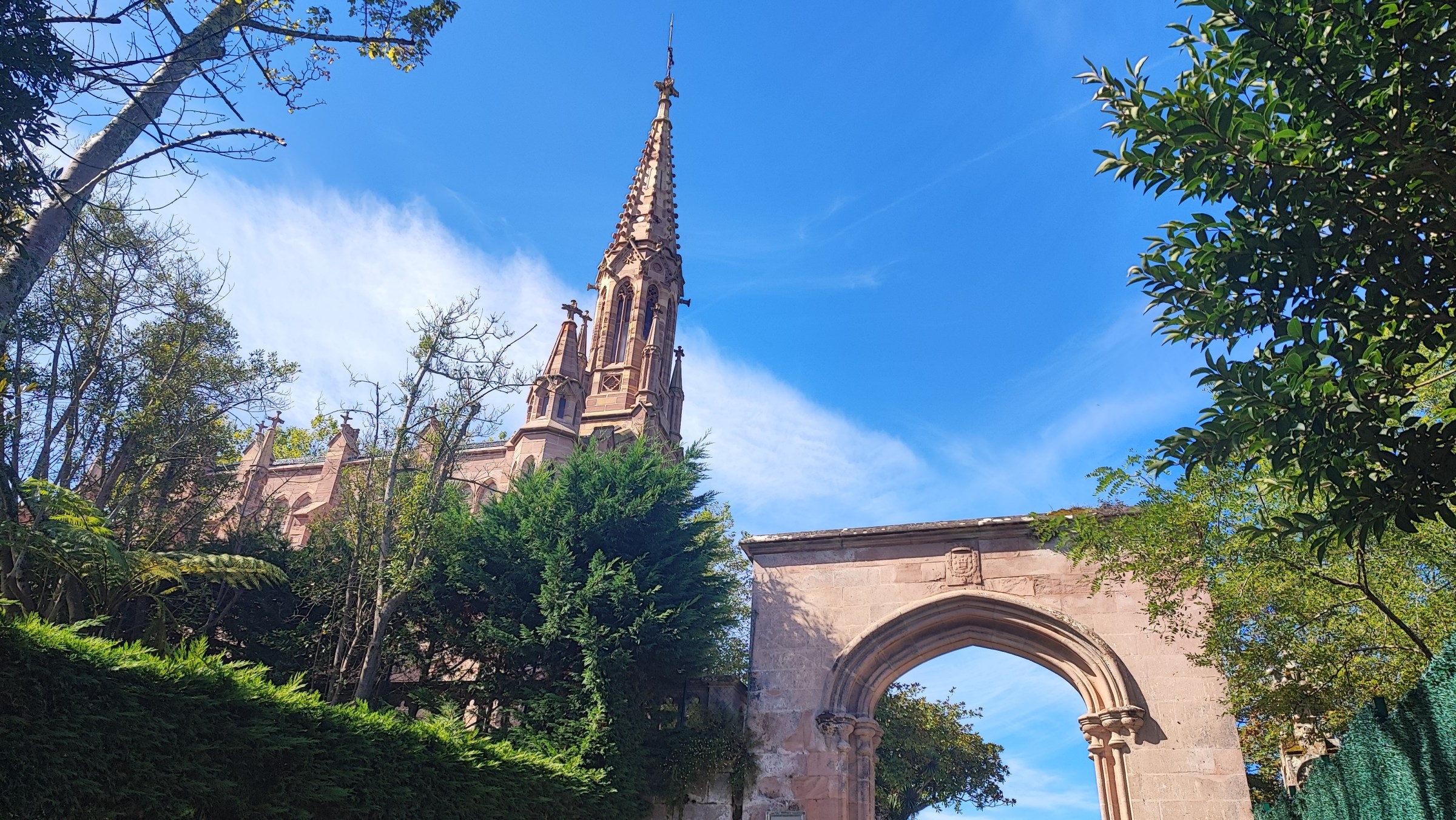 Gothic-style church spire with trees and archway under a blue sky.