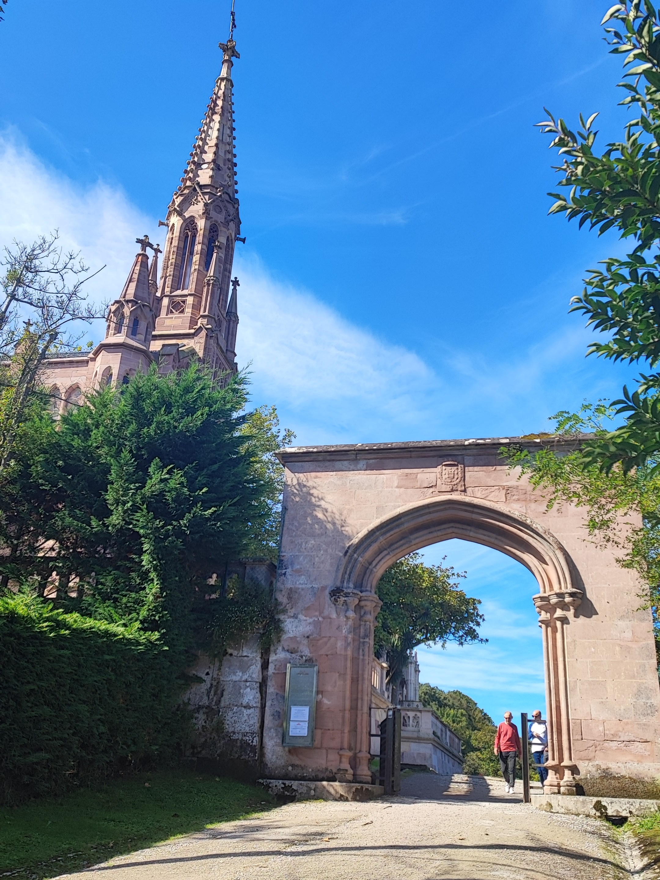 Stone archway and church spire under blue sky, person walking.