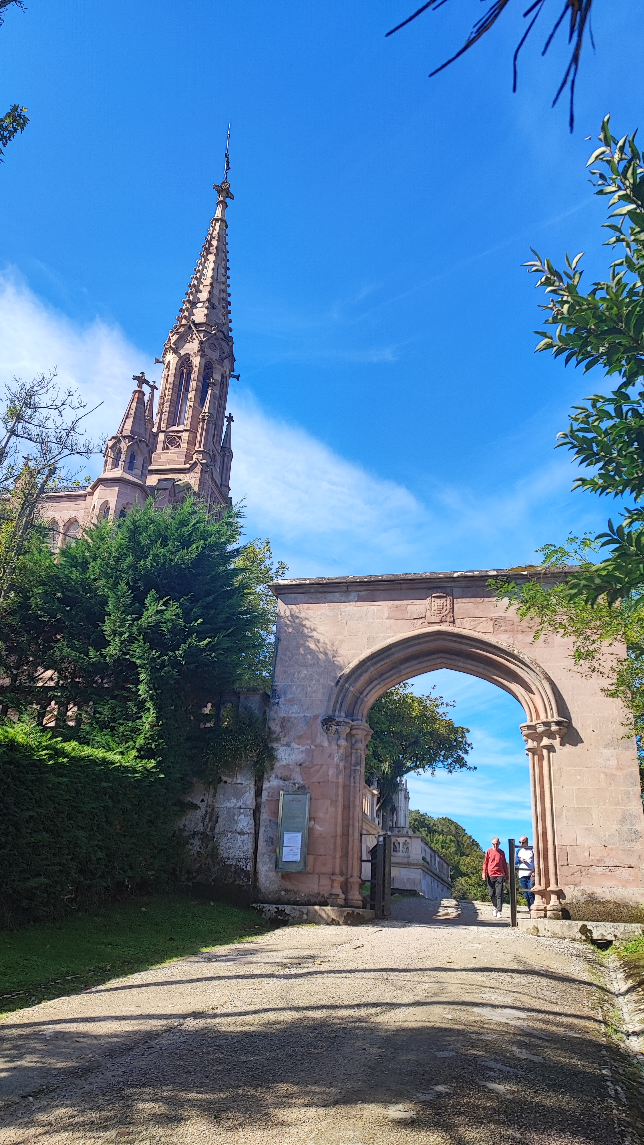 Stone archway and church spire under blue sky, person walking.