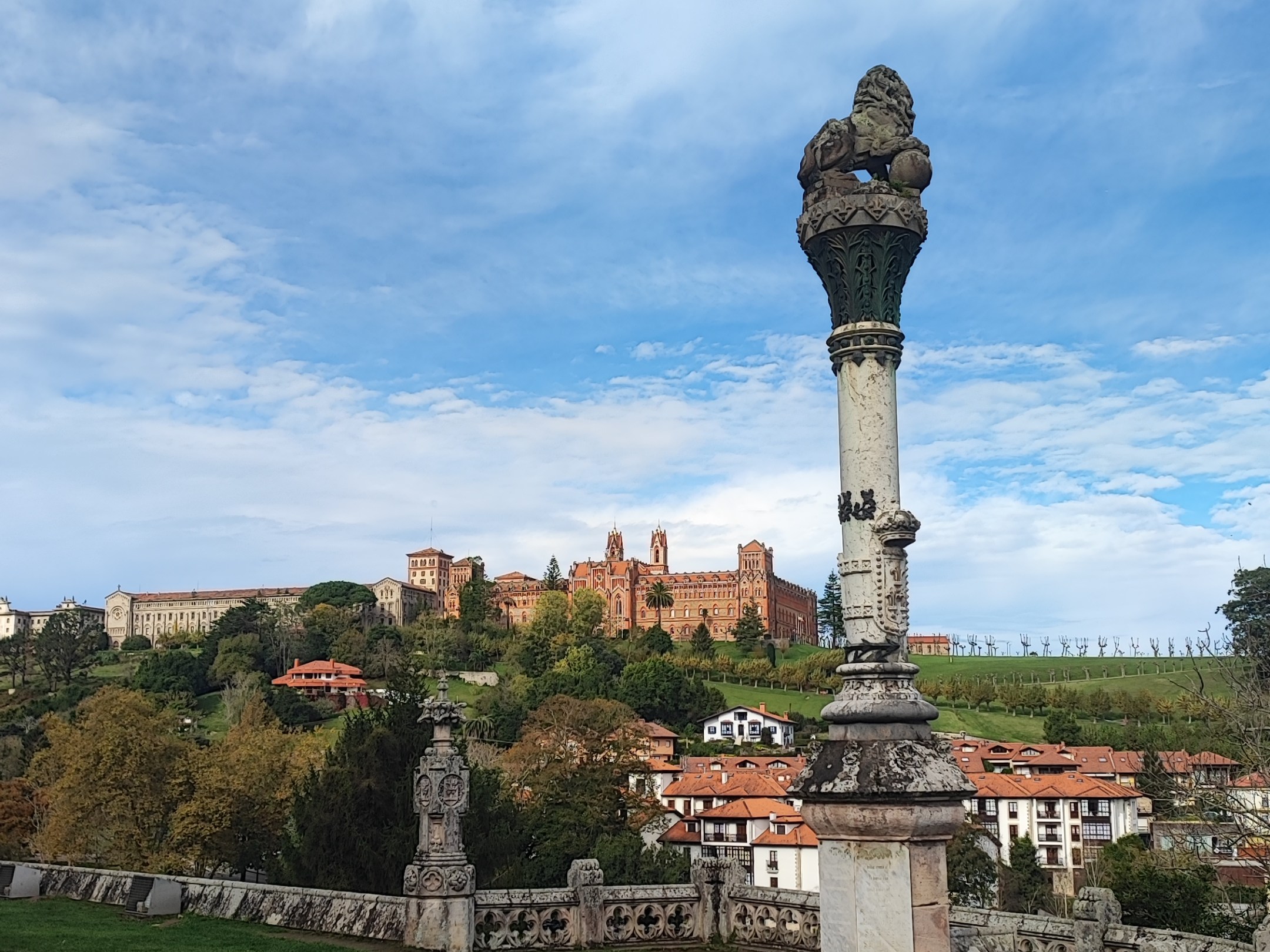 Stone column and statues with castle in the distance under a cloudy blue sky.