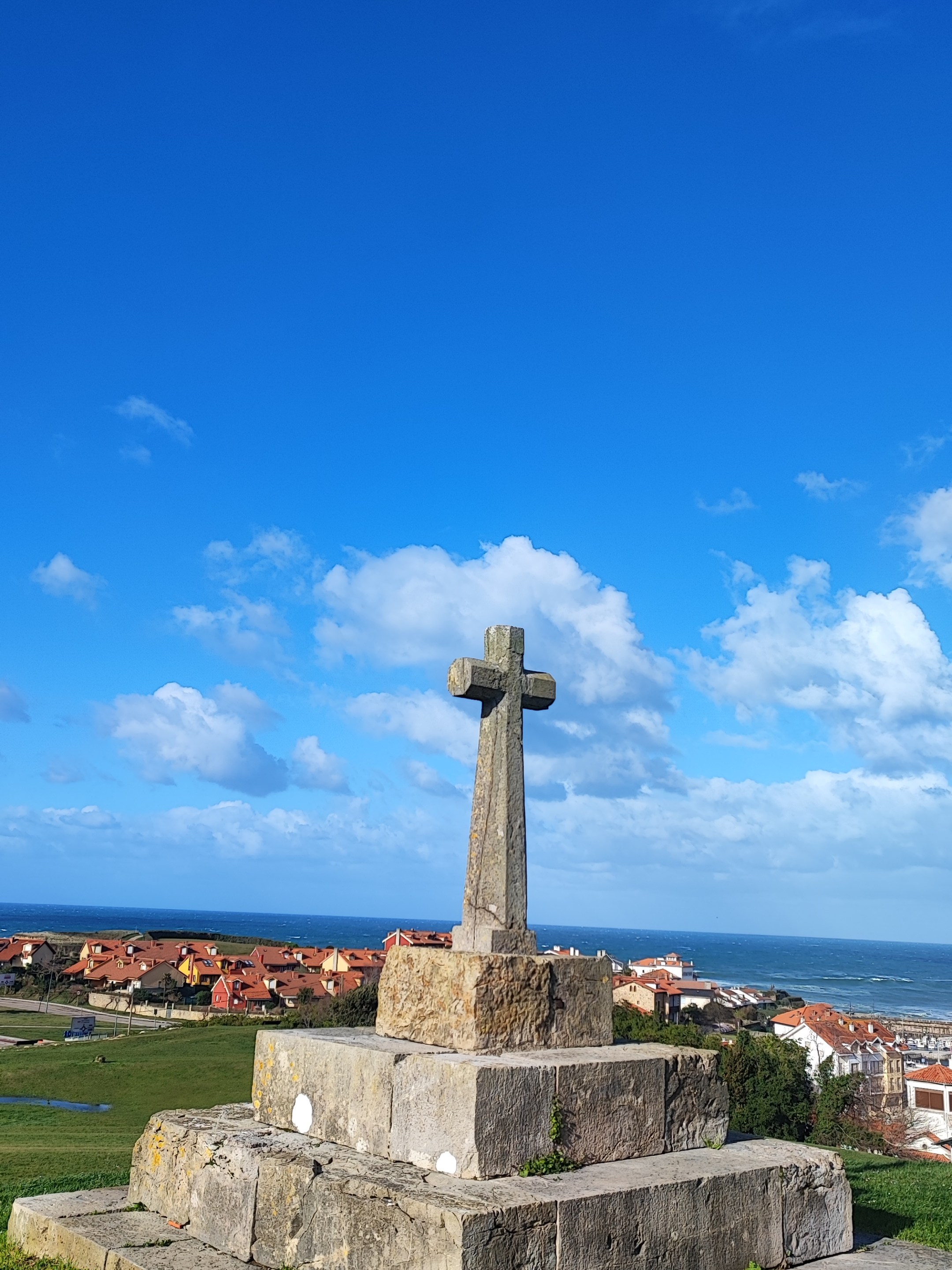 Stone cross monument on grassy hill with ocean and houses in the background.