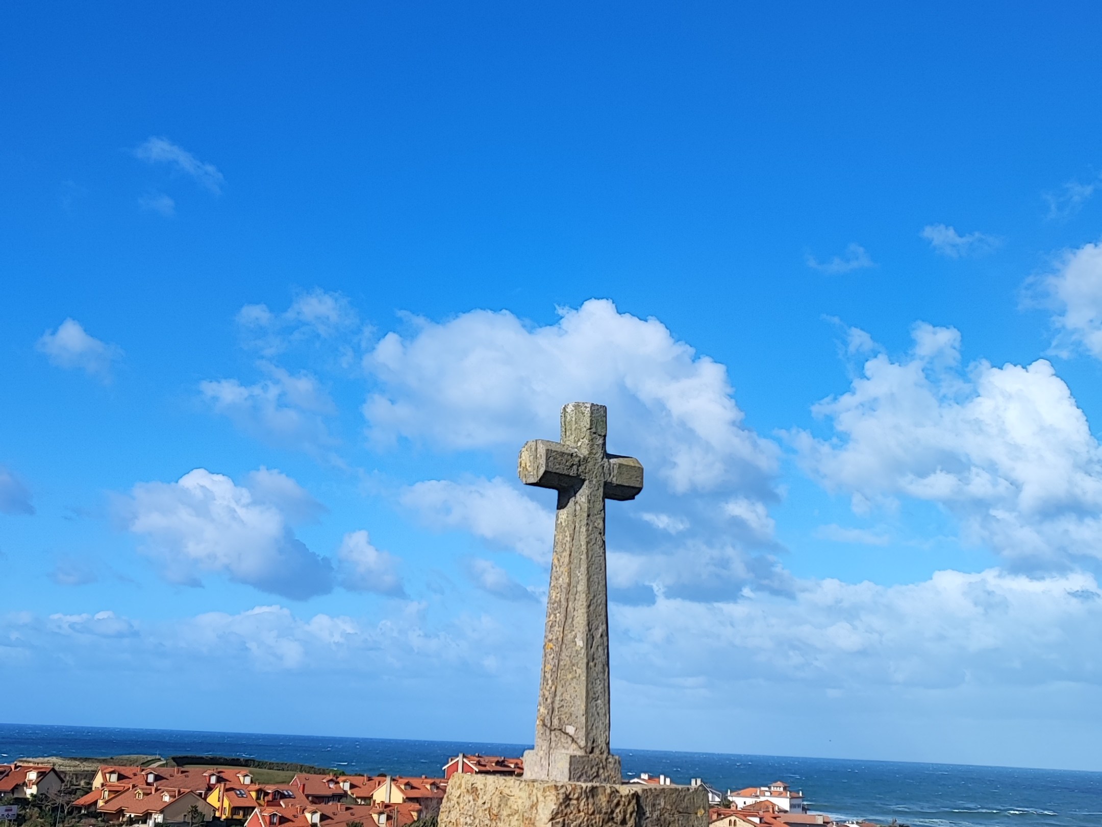 Stone cross monument on grassy hill with ocean and houses in the background.