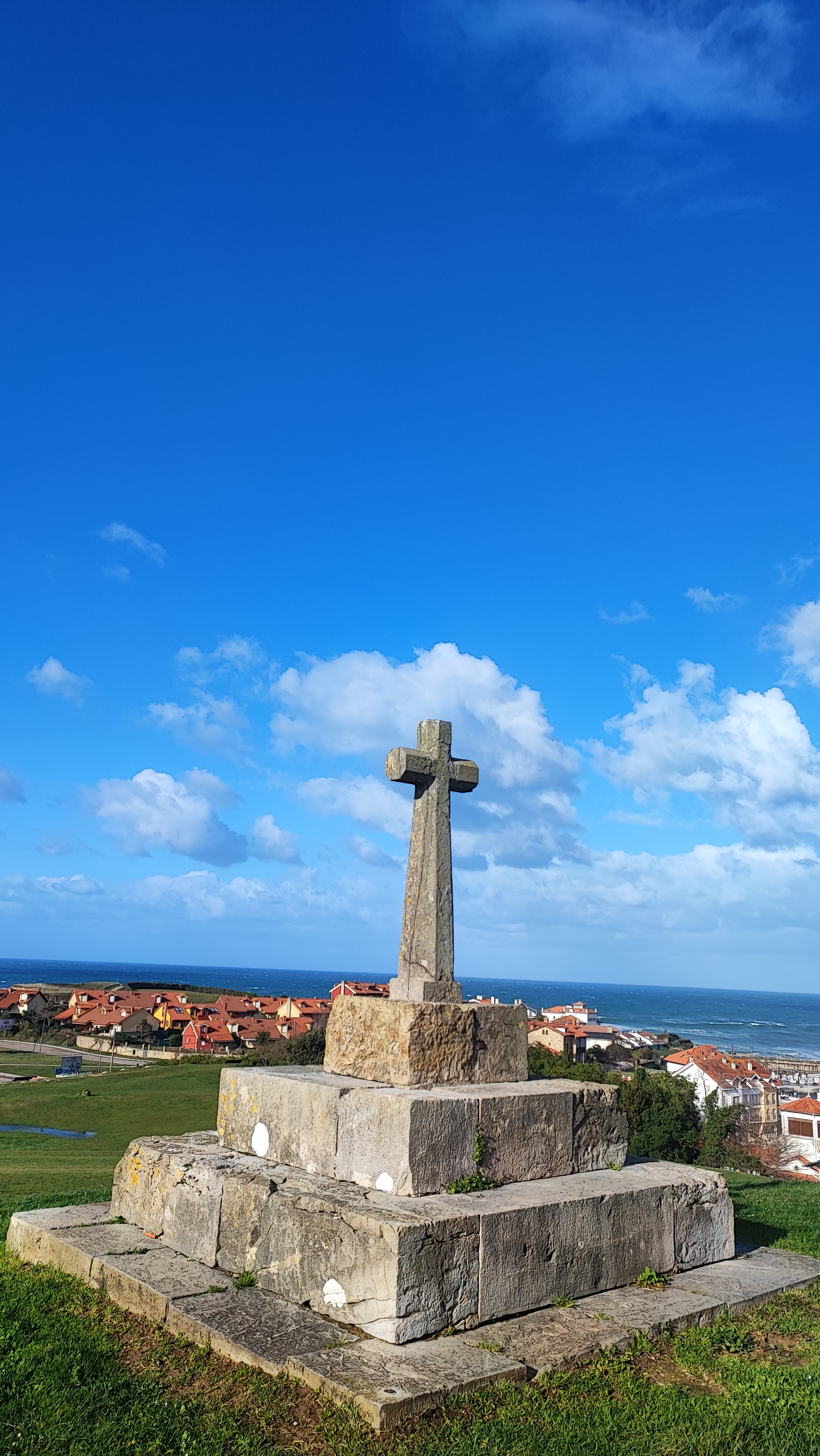 Stone cross monument on grassy hill with ocean and houses in the background.