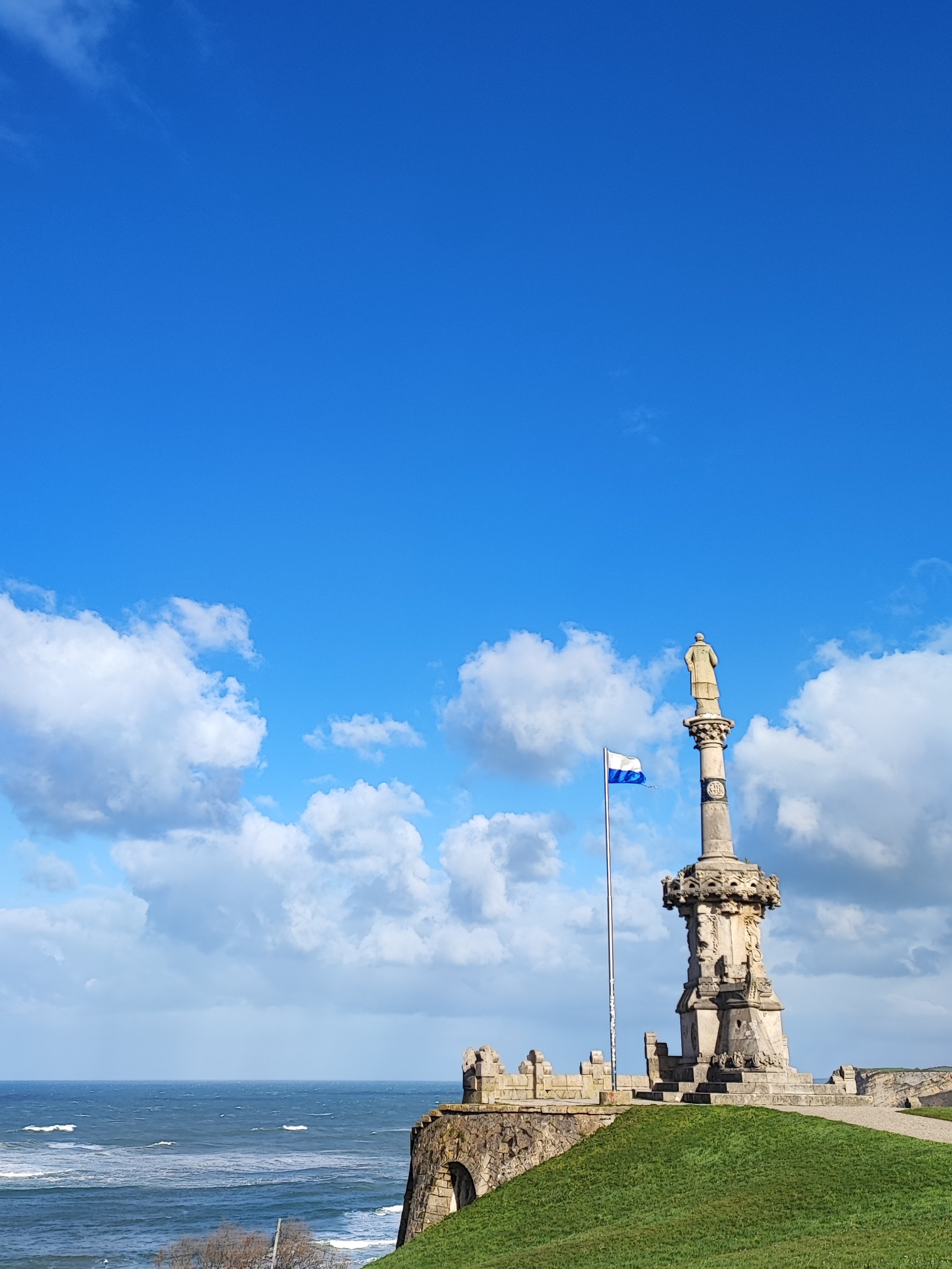 Stone monument and flag on grassy hill by the sea under blue sky.