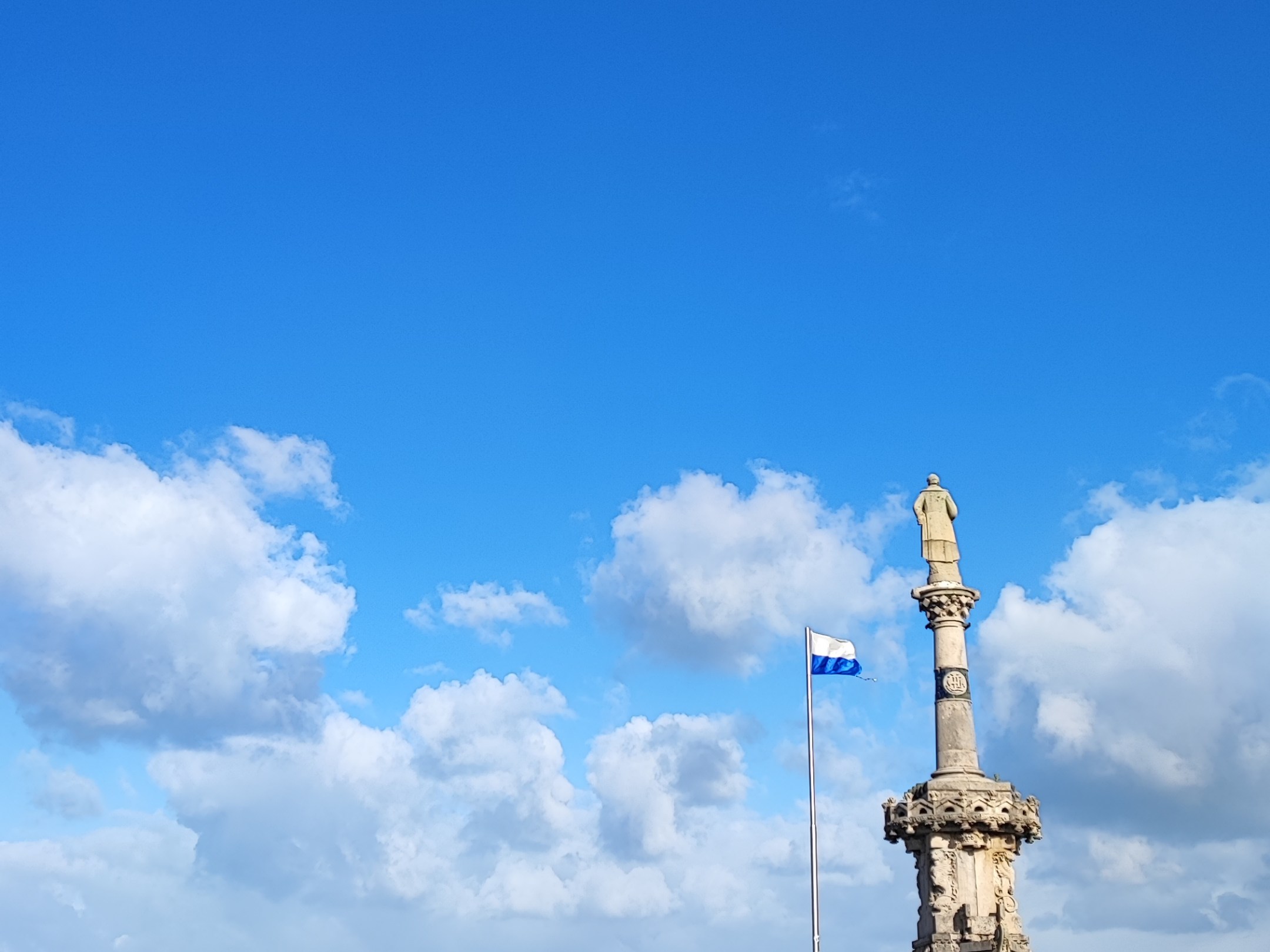 Stone monument and flag on grassy hill by the sea under blue sky.