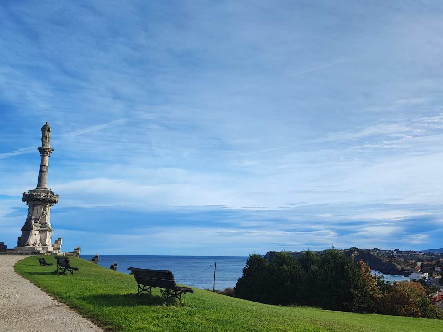 Stone monument on grassy hill overlooking a coastal town and ocean under a blue sky.
