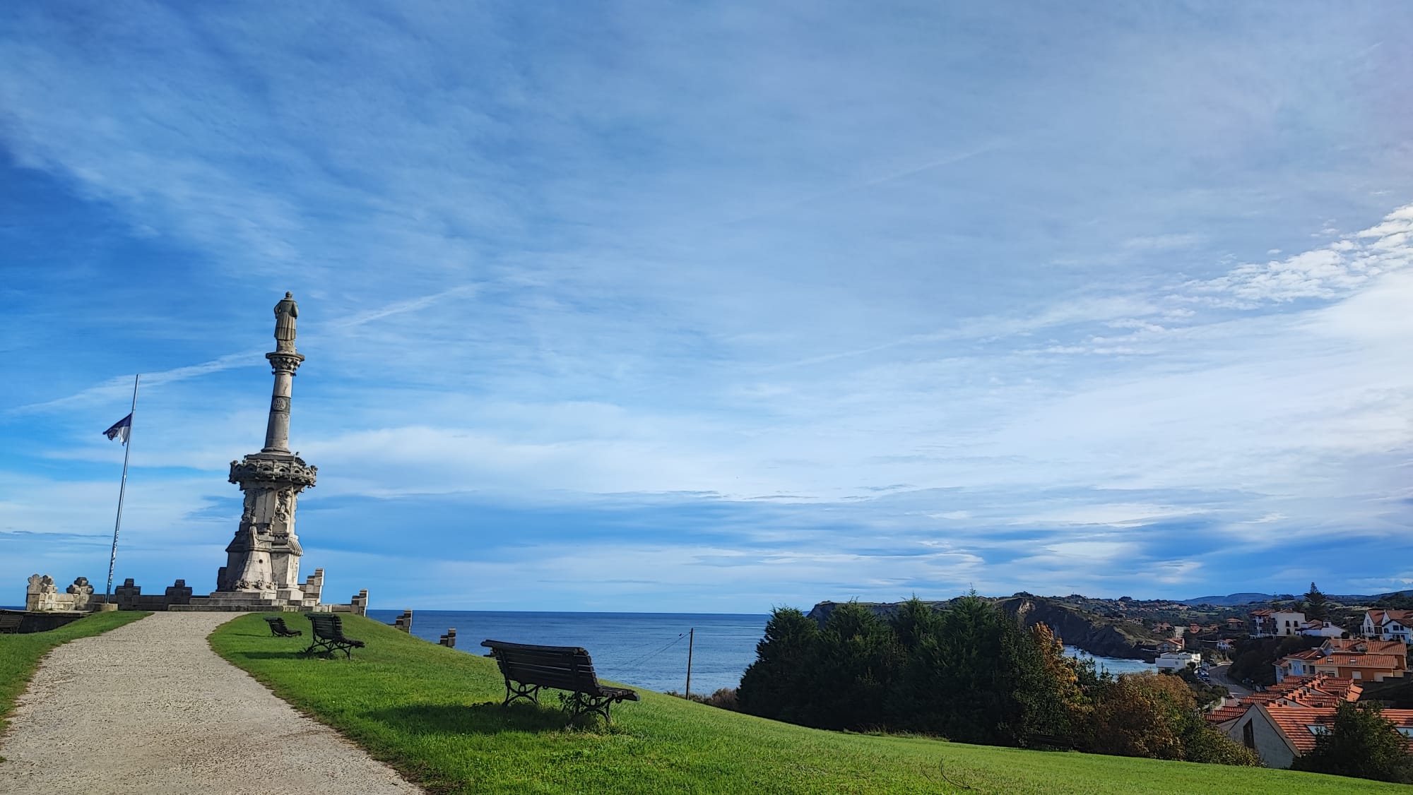 Stone monument on grassy hill overlooking a coastal town and ocean under a blue sky.