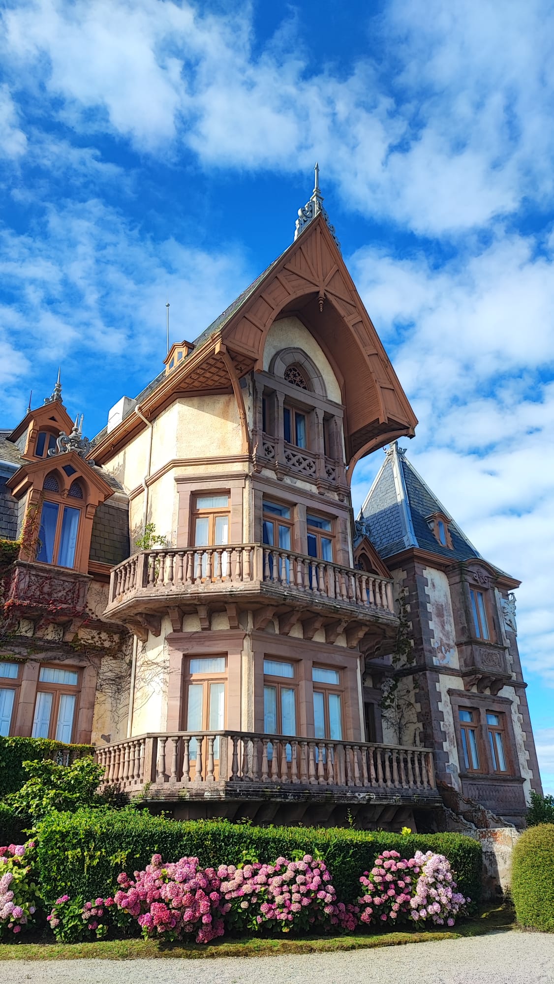 Ornate building with pointed roofs, wooden balconies, and flowered bushes under a blue sky.