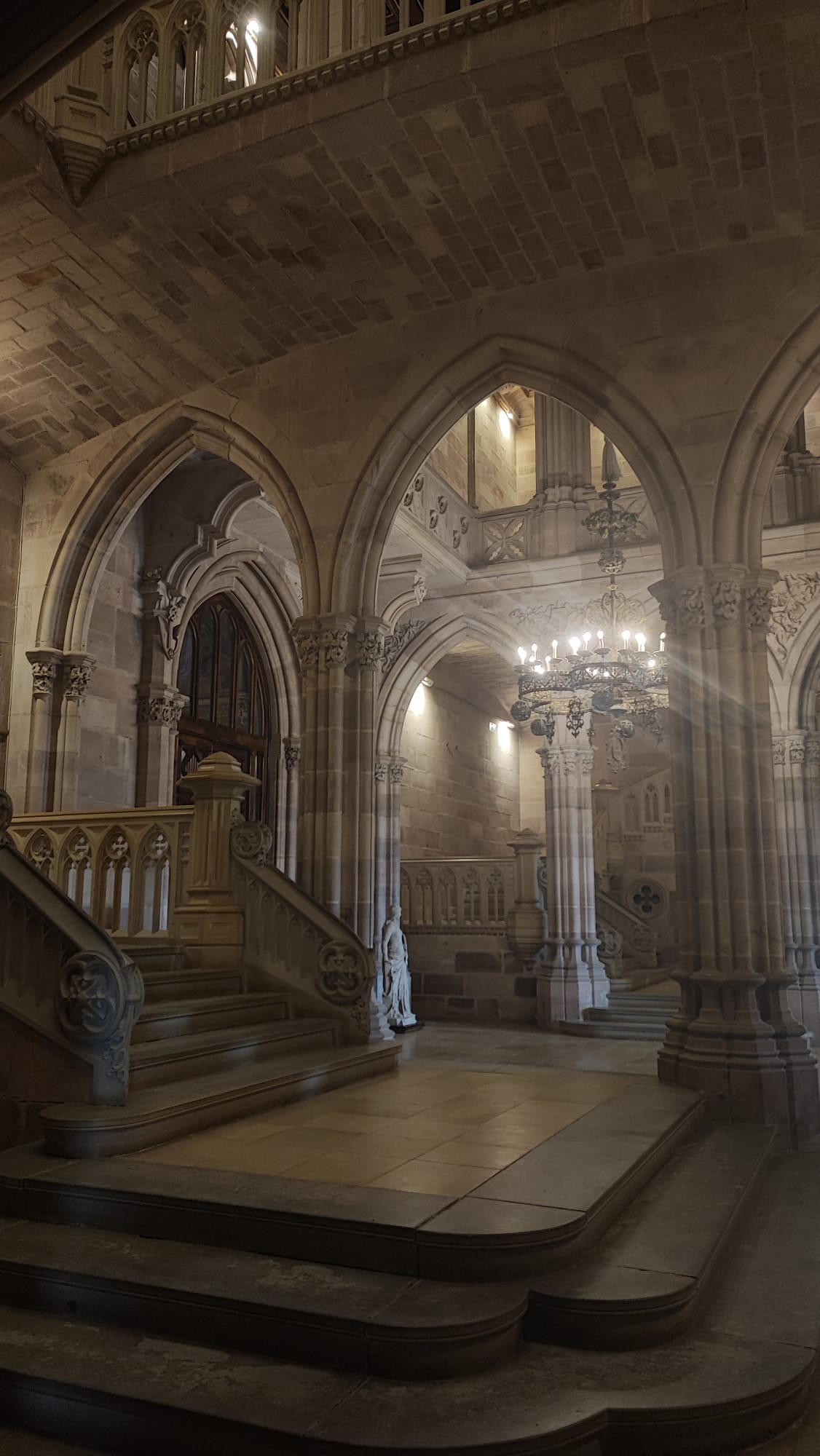 Grand stone staircase with arches and ornate chandeliers in a historic building.